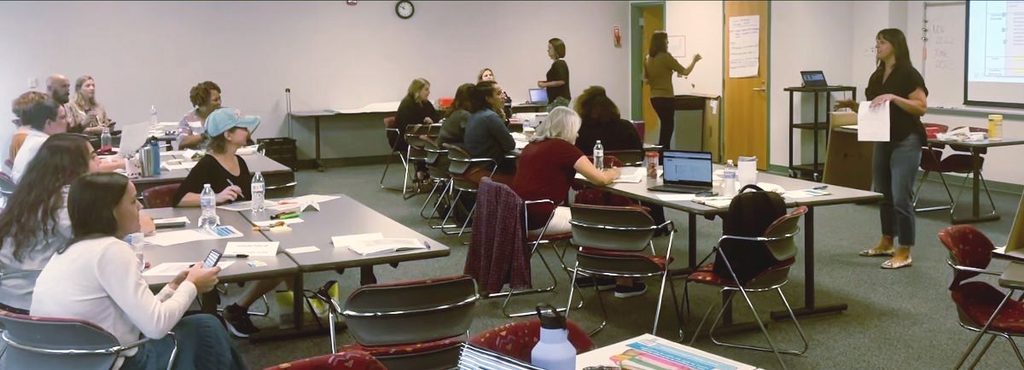 People sitting at tables with papers, facing the front of the room where a woman is talking.